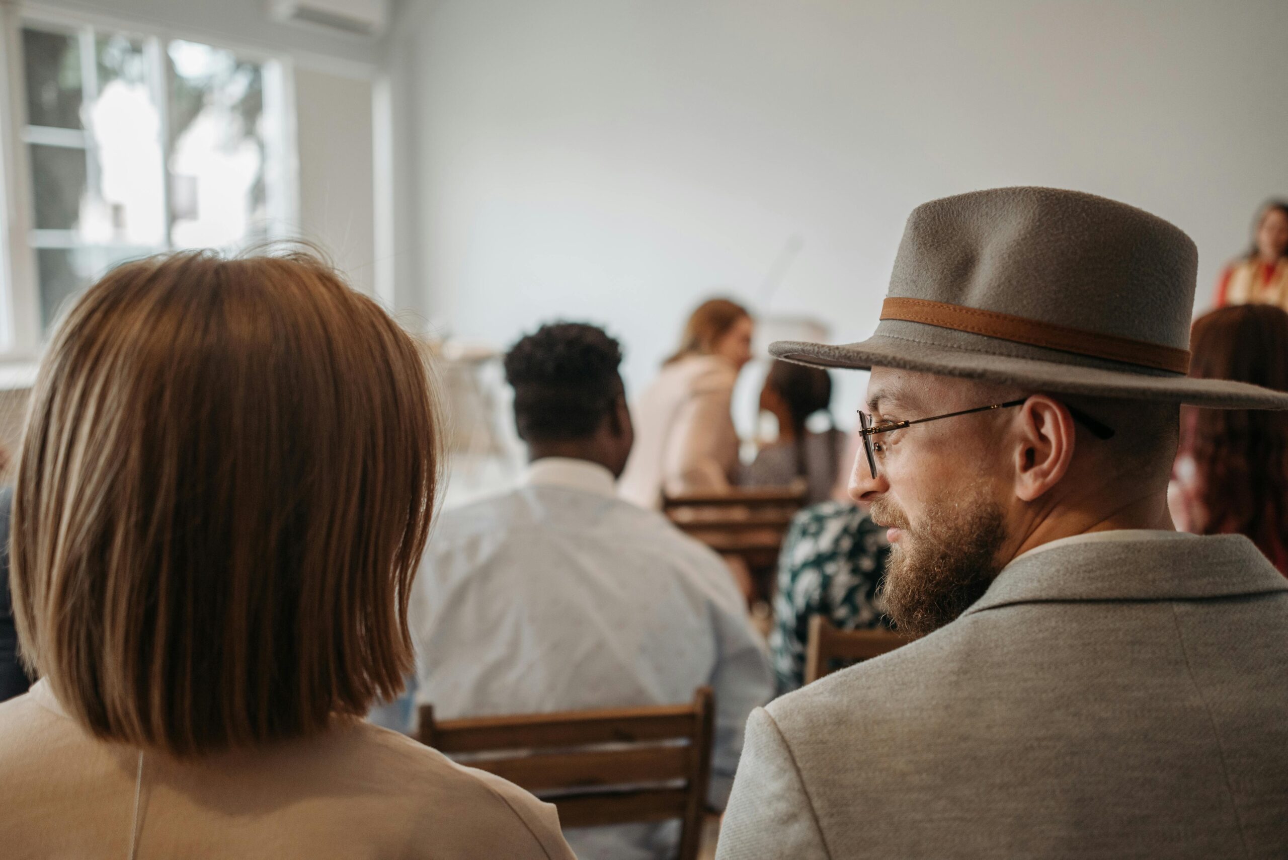 A man wearing a hat and glasses sits among attendees at an indoor seminar.